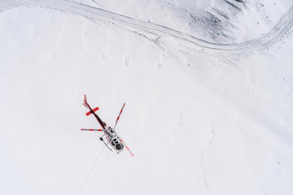 The image shows a helicopter flying over a snow-covered landscape. The helicopter has red accents and is seen from above, highlighting its spinning rotor blades. There are tracks in the snow, possibly from vehicles or machinery, visible in the top right corner of the image.