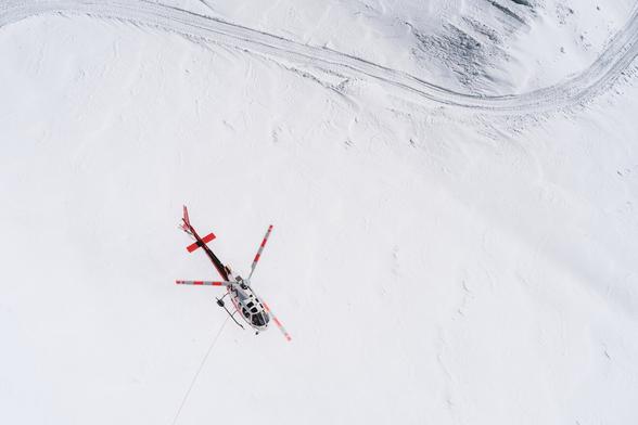 The image shows a helicopter flying over a snow-covered landscape. The helicopter has red accents and is seen from above, highlighting its spinning rotor blades. There are tracks in the snow, possibly from vehicles or machinery, visible in the top right corner of the image.