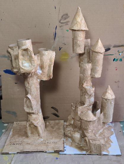 An in progress shot of two magical castles being built from card and loo roll, on a messy desk.