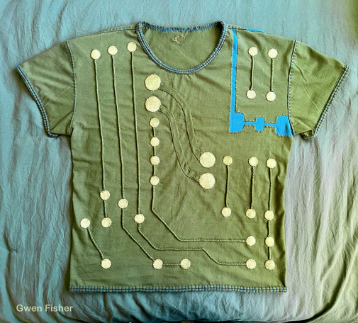 Photo of a t-shirt embroidered with a circuit design. The shoulder has a section added that looks like it’s a separate board that has been snapped apart along a line of perforation. The shirt is covered in gold circles that are appliquéd onto the shirt. The circles are connected with lines of couching, couched strips of fabric. Everything is hand sewn. The neck line and all hems are finished with the same double line appliqué stitch treatment.