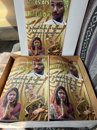 A box containing multiple copies of a book titled "Ancestors of the Rainbow: A Spectrum of Gender Before Colonization." The cover features images of diverse individuals presenting various gender identities and cultural attire.