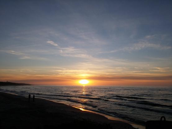 The yellow-orange sun setting over the sea viewed from the beach which is already covered in shadow. The surface of the water and the gentle sea waves reflect the sun's rays. The pale blue sky is mostly clear, with a few clear smudged clouds above the setting sun.