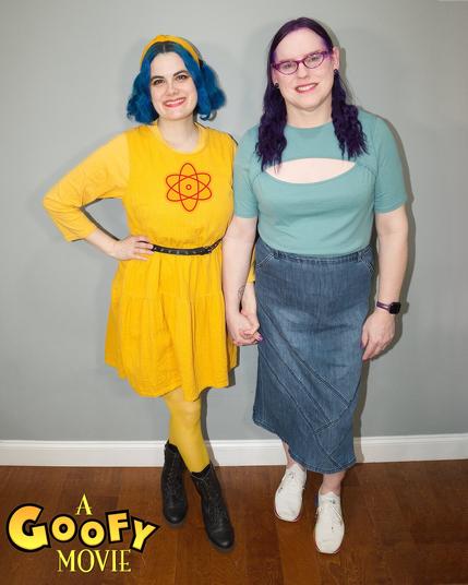 The image features two women standing side-by-side against a gray wall, with the title "A Goofy Movie" subtly integrated into the bottom-left corner.

The woman on the left has striking blue hair and is dressed in a vibrant yellow dress adorned with a red flower emblem. She confidently places her right hand on her hip while wearing black boots. In contrast, the woman on the right sports long dark purple hair and wears a light green top paired with a denim skirt and white sneakers.

The background of the image consists of a gray wall and a brown wooden floor, providing a neutral yet visually appealing backdrop for the two women. The overall atmosphere exudes a sense of playfulness and camaraderie, as if the women are about to embark on an exciting adventure together.