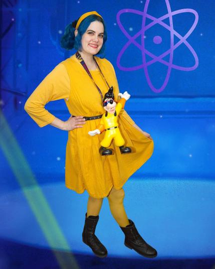The image features a woman dressed in a vibrant yellow outfit, posing alongside a small figurine that bears a striking resemblance to her attire.

Standing against a blue backdrop adorned with purple stars and an atom symbol, the woman sports a bright yellow dress paired with matching tights. Her hair is styled in two pigtails, each secured with a yellow headband. A black belt cinches at her waist, adding a touch of contrast to her outfit. The figurine, also clad in yellow, mirrors the woman's attire and hairstyle.

The overall atmosphere of the image suggests that it was captured during an event or gathering where attendees were encouraged to dress up in costumes inspired by science fiction or fantasy themes.
