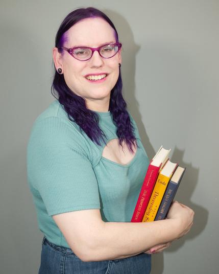 The image features a woman standing against a plain gray wall, holding three books close to her chest. She is dressed in a teal short-sleeved shirt and blue jeans.

The woman's dark hair is styled in two braids that fall over her shoulders. Her attire suggests she may be a student or someone who values education.

The books are stacked vertically with their spines facing outward, showcasing different colors: red, yellow, and black. The covers feature illegible text, but the titles "THE" and "THE" are visible on the top two books.

The background is a plain gray wall, providing a neutral backdrop for the subject. Overall, the image presents a simple yet intriguing scene that invites closer examination.