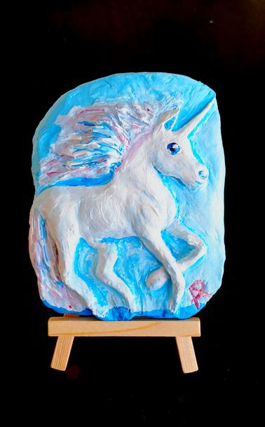 Aus Keramikmasse ein weißer, galoppierender Schimmel mit einem Horn auf der Stirn. Hintergrund und Augen sind Himmelblau und die Mähne schimmert leicht Roséfarben.