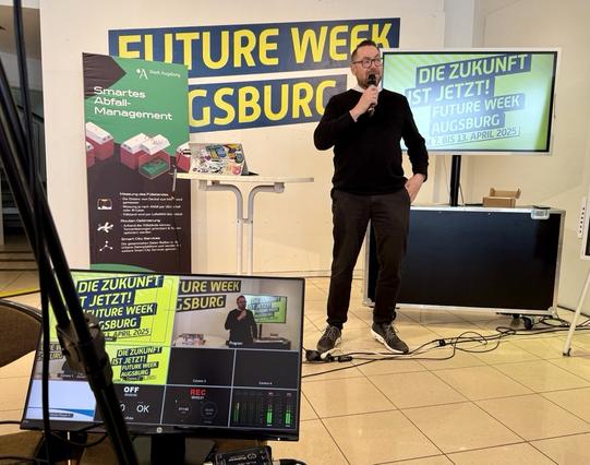 Horst Thieme, ein Mann, steht mit Mikrofon auf einer kleinen Bühne und spricht zu einem unsichtbaren Publikum. Hinter ihm befindet sich ein Bildschirm mit der Aufschrift: “DIE ZUKUNFT IST JETZT! FUTURE WEEK AUGSBURG, 7. bis 13. April 2025”. Links steht ein Roll-Up mit der Überschrift “Smartes Abfall-Management – Stadt Augsburg”. Auf einem Stehtisch liegt ein bunt beklebter Laptop. Im Vordergrund ist ein Kontrollmonitor für Videoaufnahmen zu sehen, auf dem das Bühnengeschehen live übertragen wird.