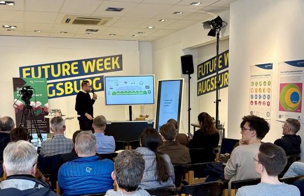 Ein Redner spricht mit Mikrofon vor einem sitzenden Publikum. Er zeigt auf eine Präsentation auf einem großen Bildschirm, die eine schematische Darstellung zur Urbanen Daten Plattform (UDP) zeigt. Im Raum hängen Poster zu Augsburger Nachhaltigkeitszielen. Die Wand ist mehrfach mit dem Logo „Future Week Augsburg“ beklebt. Eine zusätzliche Stele zeigt eine Bildschirmmaske zur digitalen Ausweisprüfung.