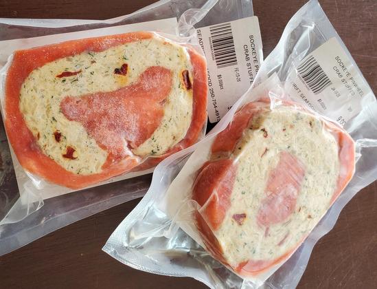 Two packages of frozen Sockeye Pinwheel Crab Stuffed seafood are featured on a brown surface. Each package contains a pinwheel-shaped portion of salmon wrapped around crab stuffing, sealed in clear plastic with labels displaying the product name, price ($9.00/pcs) & a barcode. The labels are from SEADRIFT SEAFOOD.