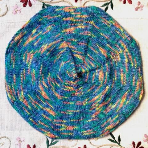 Béret tricoté multicolore à dominante bleue et verte sur un fond écru. On perçoit en haut et en bas  des broeries.