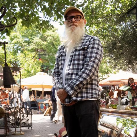 A bearded man wearing a checkered shirt, white t-shirt, and cap stands outdoors at a bustling antique market in Athens, Greece, surrounded by trees, umbrellas, and vintage items.