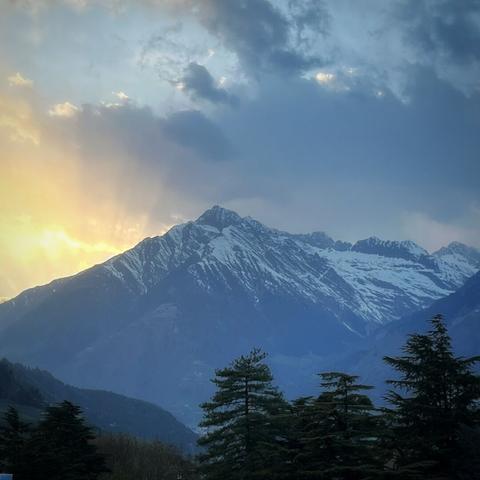 A towering mountain range dominates the center of the image, its rugged slopes partially covered with snow. The tallest peak rises sharply toward the sky. In the foreground, several tall evergreen trees create a dark silhouette against the lighter background. On the left side, the sun is either setting or rising, casting a golden glow that breaks through thick, scattered clouds. This warm light contrasts with the cool blue tones of the sky and mountains, giving the scene a dramatic and serene atmosphere. The valley below is shadowed and faintly visible, suggesting depth and scale.
