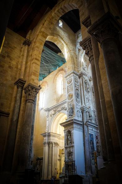 The image depicts the interior of a grand cathedral, showcasing its architectural beauty. The foreground features tall, ornate columns with intricate carvings, leading the eye towards the central archway. The archway is adorned with detailed sculptures and reliefs, enhancing the sense of grandeur. The walls are made of light-colored stone, with some areas showing signs of wear, adding to the historical ambiance. Sunlight filters through the windows, casting a warm glow and creating a contrast between light and shadow. The ceiling is supported by a network of beams, and a green protective netting is visible, suggesting ongoing restoration work. The overall atmosphere is one of reverence and historical significance.

Provided by @altbot, generated privately and locally using Ovis2-8B