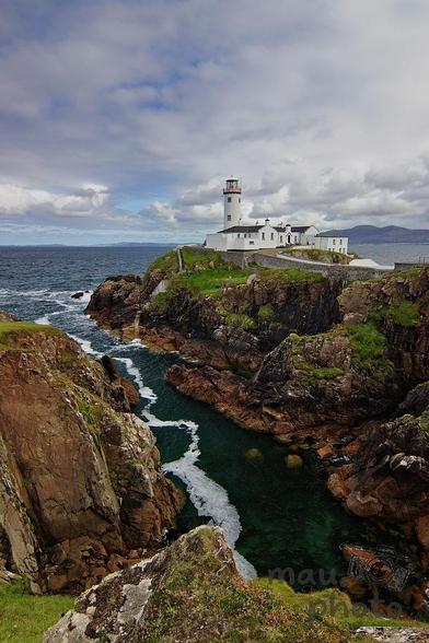 The image shows a scenic view of a lighthouse situated on a rocky cliff by the sea. The lighthouse is surrounded by white buildings and a stone wall. The area is lush with green grass, and the sea below has crisp, clear water. The sky is partly cloudy, adding to the dramatic landscape.
