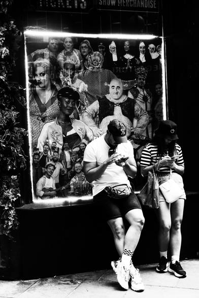 Black and white. A young man and woman, probably in their twenties, dressed like London tourists in the sunshine, in shorts, t-shirts, and a baseball cap for him and a bucket hat for her, leaning against a large poster, hunched over their phones. The poster is an image of characters from London shows. The only character recognisable to me is Shrek. The entire show of real life is going on around the couple, but it's all filtered out for them apart from a few inches of screen space.