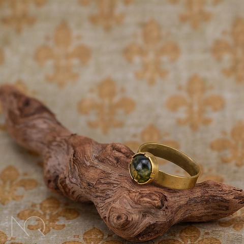 En una rama nudosa en primer plano descansa un anillo de latón sencillo, solo una banda plana y lisa y como adorno un cabuchón redondo de 6mm de una ágata musgosa sintética