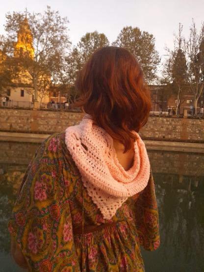 Yo con un vestido fino primaveral frente a un río y de espaldas a la cámara, con un fular de crochet a los hombros.