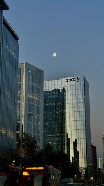 A low-angle view of several modern glass skyscrapers at dusk, with a bright full moon visible in the twilight sky.  A red city bus is partially visible in the foreground. The tallest building displays the logo for a company called "Sura".  Trees are partially visible between the buildings.