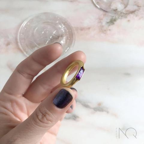 una m ano sujeta un anillo  dorado de perfil redondo con una piedra tallada morada y rectangular