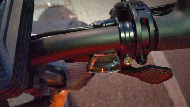 Nahaufnahme der rechten Lenkerhälfte meines Brompton Electric G-Lines. Unter dem Griff sieht man prominent einen Schalthebel für den Daumen. Links daneben ist ein Ganganzeige hinten einem Fensterchen in Bogenausschnittsform. Der hellrote Zeiger steht ganz am linken Rand auf "4". Knapp links der Mitte leuchtet ein senkrechter, roter Streifen an der Vorderkante des Fensters. Es ist die Spiegelung einer auch noch zur Seite leuchtenden Rückleuchte am Sattelrohr.