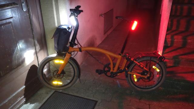 Mein Brompton Electric G-Line in momentaner Vollbeleuchtung: Hochkant montierte, weisse bzw. rote Akkuleuchten an Sattelstange und Lenker und die vom E-Bike-Akku gespeisten Leuchten ab Werk zwischen Kotflügel und Fronttasche und hinten am Gepäckträger. Ausserdem leuchten noch die zwei Paar gelbe Speichenreflektoren durch die Aufnahme mit Blitz.
