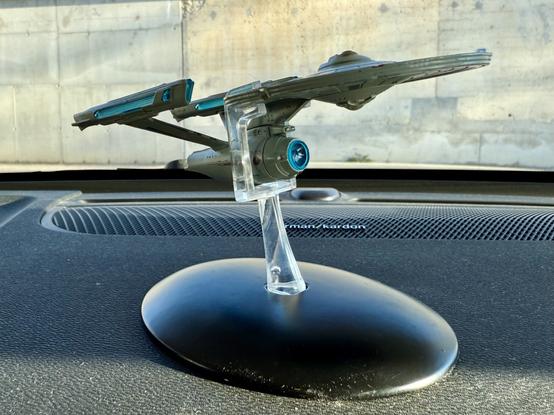A model spaceship displayed on a car dashboard. The spaceship is mounted on a clear stand that sits on a black base. The background features a textured surface, possibly a wall.