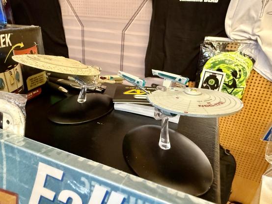 A display of two model starships from the "Star Trek" franchise, each prominently featured on black bases. Various items such as books, pens, and merchandise are visible in the background.