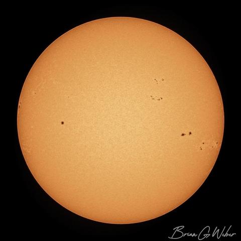 A colorized orange solar image with the full disk of the sun dotted with sunspots mostly clustered on the right side