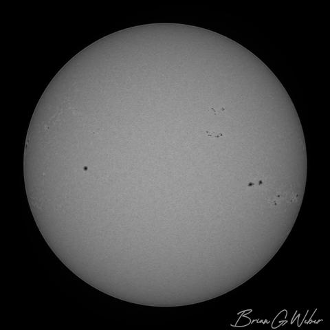 A monochrome solar image with the full disk of the sun dotted with sunspots mostly clustered on the right side