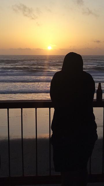 A person in silhouette leans against a railing, watching a sunset over the ocean. Waves can be seen in the foreground, and a bottle is placed on the railing next to them. The sky is illuminated with warm hues of orange and yellow.