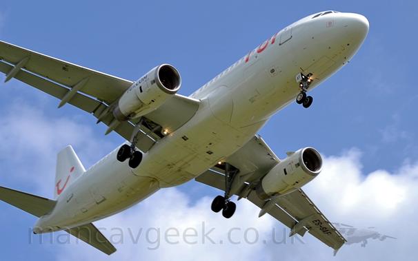 View from almost underneath of a white, twin engined jet airliner flying from left to right at a very low altitude, with undercarriage lowered and flaps extended from behind the wings, suggesting it is about to land.
The plane is pretty much just white, with red "TUI" titles on the upper forward fuselage, and a red logo that looks like the upper-case letters "T", "U", and "I" smushed together to form a winking smiley face.
There are black "Tui[dot]com" titles on the outer faces of the engine pods slung under the wings.
The registration "ES-SAF" is under the outer left wing.
Bright lights in the landing gear wells are shining forwards.
Clouds fill the background under the plane, with blue sky above.