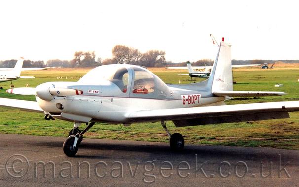 Side view of a single engined light aircraft parked facing to the left.
The planes upper surfaces are a metallic, silvery-grey, with white lower surfaces, and red piping.
The registration "G-BOPT" is on the rear fuselage in red.
The tail is white, with a grey leading edge, and a red beacon on the top.
A large grass airfield fills the background, leading up to trees in the distance, with a couple of other light aircraft parked on the grass.
Bright but hazy sky fills the rest of the frame.