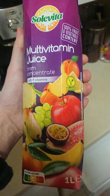 A box of 1L (Lidl) Solevita multivitamin juice. Half of the box is purple with the rest xovered by photos of fruits. Part of the hand holding them, is in the frame. Photo is taken in a kitchen environment.