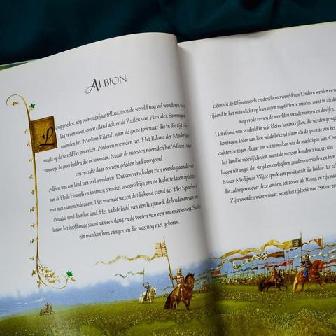 A book lying open with an illustration that covers the bottom of both pages. It shows many knights on horseback carrying banners of arms. The banners are long and trailing behind them. The text above it starts with an illuminated letter.