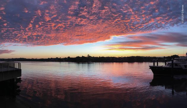 A spectacular sunrise over Brisbane river. The dark silhouette of the opposite river bank splits the image. Overhead, a uniform bank of rough-textured cloud is lit blazing red-pink by the not-risen sun, the undulations in the cloud base thrown into strong relief by the intense pink light. Below, a calm river reflects some of the scene above, with moored boats and a walkway bracketing the shot right and left.
