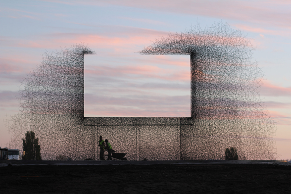 A photo of the sculpture "Non Sign II" designed by Seattle artists Annie Han and Daniel Mihalyo. The work resembles a cloud of wires or steel wool, with a perfectly rectangular void cut out of the center in the size and shape of a highway billboard. In the background, wispy pink and white clouds fill a blue sunset sky. In the foreground we see the silhouette of two human figures, emphasizing the large size of the artwork.