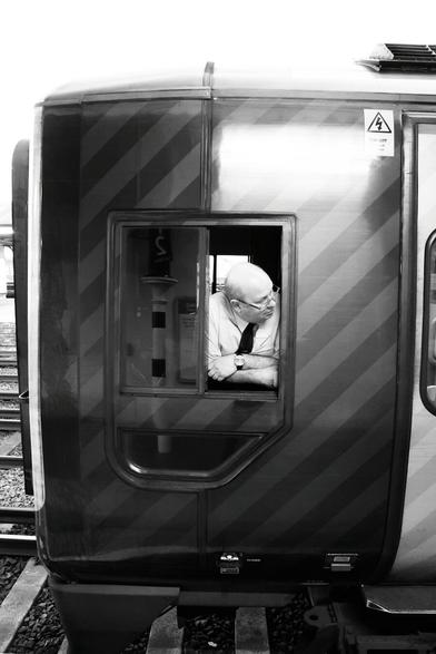 Balding train driver, with white shirt and black tie, cross-armed, leaning on the windowsill of his train at Eastleigh Station, looking out of his window to make sure everything is safe before he moves off.