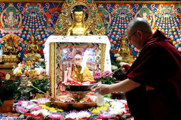 A Buddhist monk in a saffron robe and shaved head with glasses spoons water from a small fountain on a colorful altar with a photo of the Dalai Lama behind the fountain and a large Buddha statue flanked by colorful images and decorations in the background.