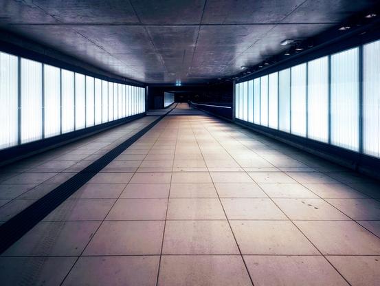 A symmetrical, eye-level shot of a long, empty underpass or walkway. The walls on either side are lined with evenly spaced, vertically rectangular light panels, emitting a bright, cool-toned light. The floor is tiled with large, square, light-coloured tiles, showing some faint marks. A dark, textured tactile paving strip runs down the centre of the walkway. The ceiling is a plain grey concrete. In the distance, the walkway curves slightly to the right and becomes darker. An illuminated green emergency exit sign is visible on the ceiling further down the walkway. The overall atmosphere is clean and somewhat sterile.