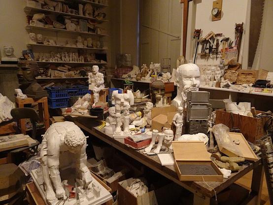 Interior de un estudio lleno de esculturas, muchas de ellas a medio realizar (bustos, cuerpos, pies), en mesas, cajas, estanterías, el suelo... Algunas herramientas en la pared.