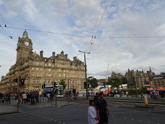 Un gran edficio histórico monumental (un hotel) en una plaza en una ciudad. En primer plano, gente caminando por la calle. Imagen atravesada por los cables del tranvía.