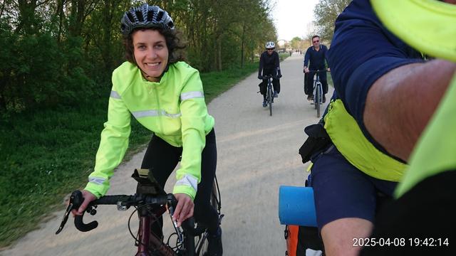 Femme au joli sourire habillée de jaune et noir sur un vélo gravel, entre un cycliste randonneur et 2 vélotafeurs  à l'arrière plan, sur une large piste cyclable entre des bois.
