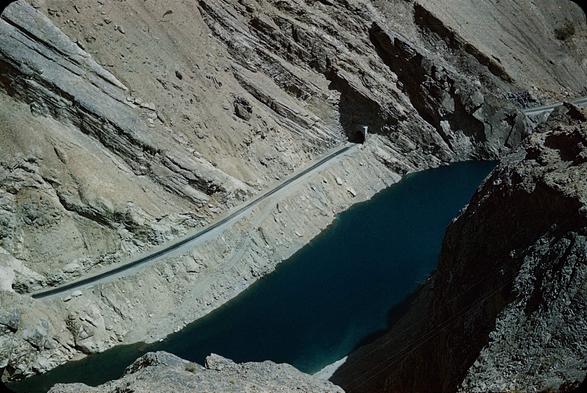 Steep sloped cliffs with rock strata at a 45 degree angle. Near the bottom is a narrow road that is heading to a tunnel that goes through some of the interesting strata. Below that is a blue green lake that is rather narrow and confined by the rock formations.