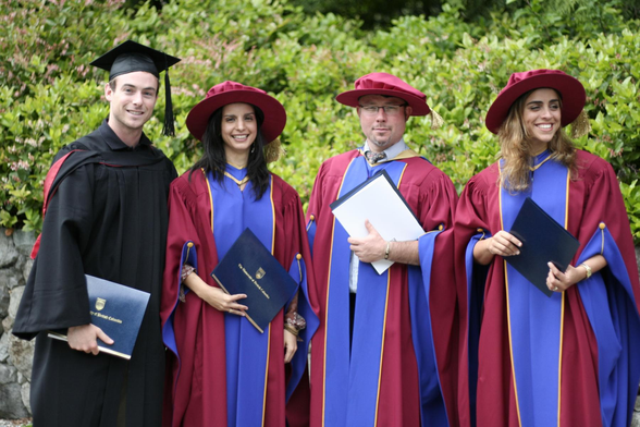 Four graduates from UBC stand holding their diplomas. The leftmost is wearing a black gown and a mortarboard, and the other three are wearing a maroon gown with blue sleeves and front along with a maroon hat.