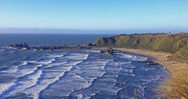 Photo shows a pebble beach in a half moon bay with deep blue waves in rows coming in from the deep blue sea to the right. And in the background behind a rocky outcrop forming a barrier north of  the bay, the blue sea is stretching far into a cloudy horizon in the distance. A steep ridge is raising up from the beach, forming a hard to climb rocky barrier towards the inland. On top of this barrier there is a rich growth of coastal flora of bushes, turf, grass, flowers and some low, windswept trees. At the far end of the cliff a lighthouse is visible at the west cape outcrop.  It is white, with roofs and details in the building painted red. Some small houses and buildings nearby are the home of the guardian/operator of the lighthouse. Even if the background cloud over the sea is overcast, the bay and land area are bathed in sharp sunlight just before it changes to warmer sunset colours.