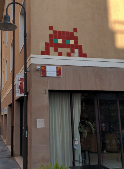 Au coin de la "via Boccaccio"', en hauteur, un grand Invader, les pixels figurés par des carreaux carrés rouges, les yeux verts et blancs.