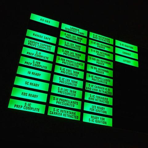 A dimly lit control panel displays various illuminated rectangular buttons, each labeled with text related to rocket launch sequences. The glowing green buttons include labels such as "AS-503," "Launch Sequence Start," "S-IVB Fuel Tank Pressurized," "Commit," and "Liftoff."