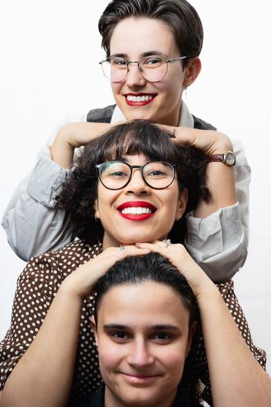 Three individuals are arranged in a vertical stack against a white background. The top person has short hair, round glasses, and a bright red smile and is lightly resting their chin on the head of the middle person. The middle person has curly, dark hair with a fringe, wears glasses and bold red lipstick, and rests their hands gently on the lower person’s head. The bottom person has their hair tied back and smiles softly. All three look toward the camera, creating a playful, tiered portrait.