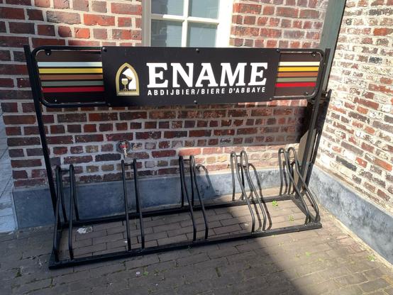 A bicycle rack. It is made of metal and has a place to hold five bikes by the front wheel. Across the top of the race is a banner sign for Ename Belgian beer. It reads:

ENAME
ABDIJBIER/BIERE D'ABBAYE