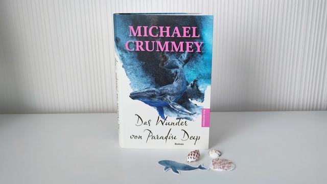 Auf einer hellen Fläche vor einer ebenso hellen Wand steht das Buch.  Rechts neben dem Buch befinden sich drei helle Muscheln mit bräunlicher Musterung sowie ein Aufkleber mit einem blauen Wal. Das Buchcover ist in einem beigefarbenen Grundton gehalten. Im unteren Bereich steht auf dem hellen Hintergrund in einer zweizeiligen, schwarzen Schmuckschrift der Titel „Das Wunder von Paradise Deep“, direkt darunter in kleiner, fetter Schrift der Zusatz „Roman“. Ganz am rechten Rand befindet sich ein rosafarbenes Rechteck mit dem Verlagsnamen in weißer Schrift. Im oberen Bereich ragt eine Illustration ins Bild, die Wellen von unten zeigt – darin schwimmt ein großer Wal. Diese Illustration ist in verschiedenen Blautönen gehalten. Am oberen Rand steht der Name des Autors in pinker Schrift mit großen Serifenbuchstaben.